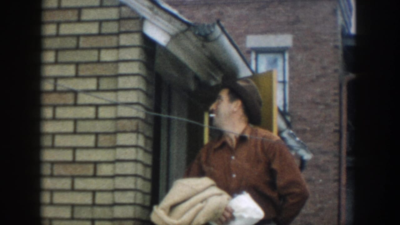 In 1957 Michigan home-movie footage, a man in a brown shirt and hat stands at a brick building entrance holding fabric and paper.