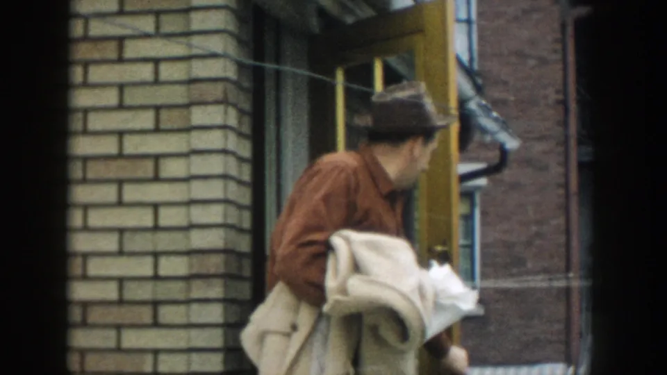 A man in mid-century clothing turns toward the building entrance, holding a blanket-like bundle beside the yellow door frame.