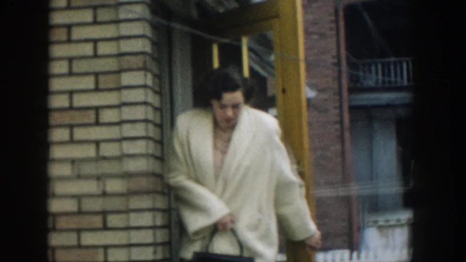 A woman in a light-colored coat steps out of the brick building doorway in the 1957 Michigan home-movie scene.
