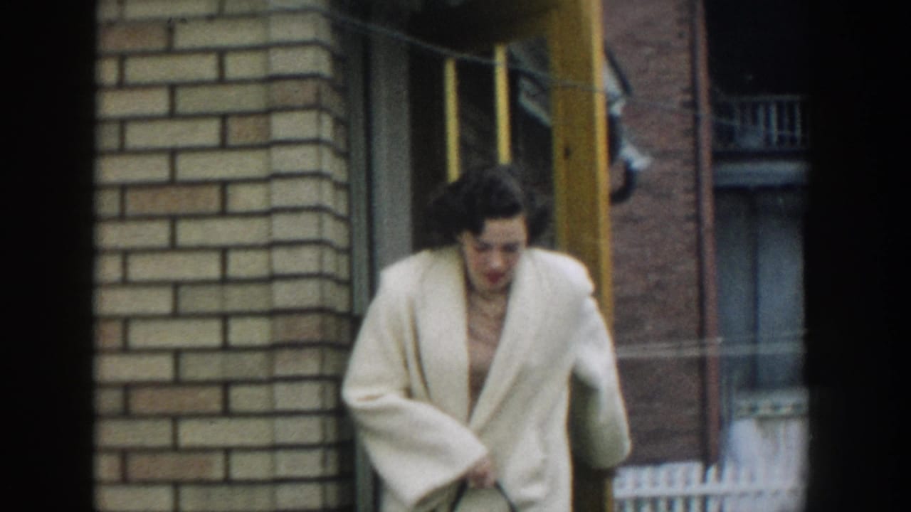 In 1957 Michigan home-movie footage, a woman in a white jacket steps out from a brick doorway and looks down as she exits.