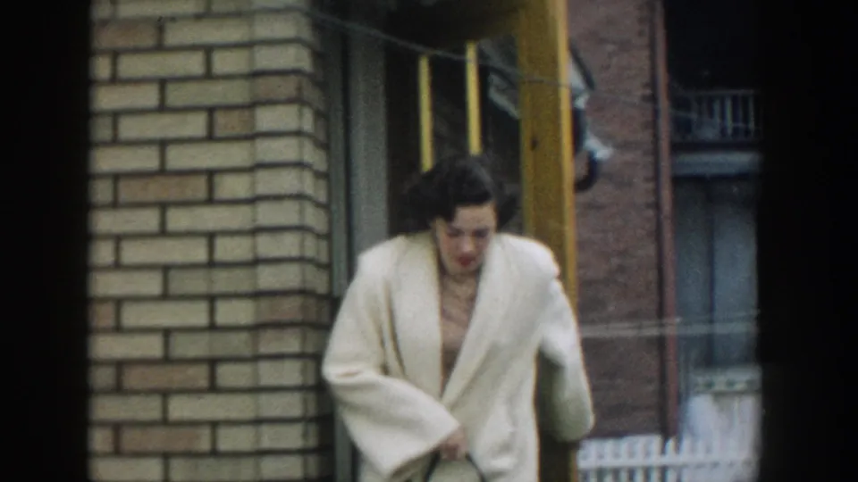 In 1957 Michigan home-movie footage, a woman in a white jacket steps out from a brick doorway and looks down as she exits.