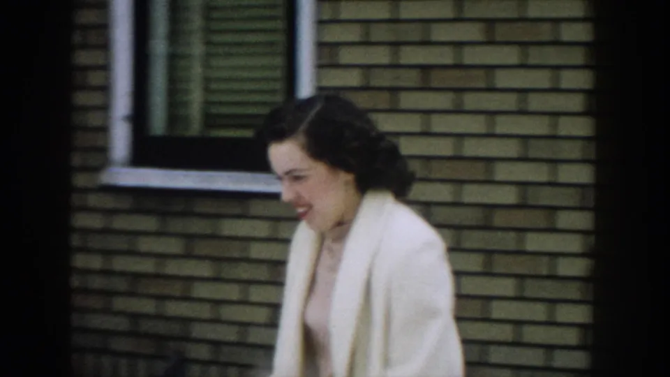The woman smiles shyly as she moves past the brick exterior, with a window and wall filling the background.