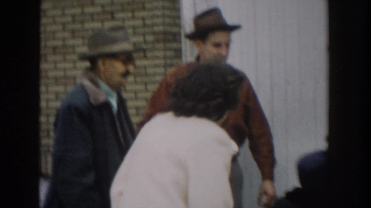 1957 Michigan 8mm frame showing adults gathered outdoors near brickwork and siding as a woman in a white coat leans toward a bundled baby.