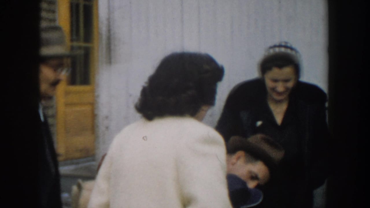 1957 Michigan home-movie frame of a woman in a white coat greeting a bundled baby beside a smiling woman in a dark coat and headband.