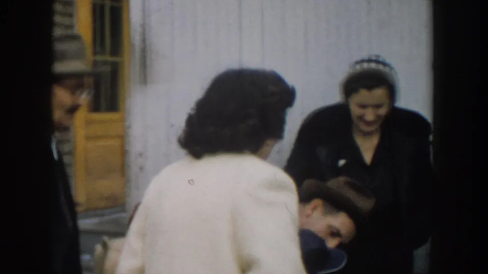 1957 Michigan home-movie frame of a woman in a white coat greeting a bundled baby beside a smiling woman in a dark coat and headband.