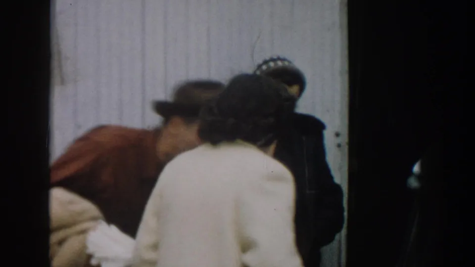1957 Michigan 8mm frame showing several adults leaning into a close family greeting beside pale exterior siding.