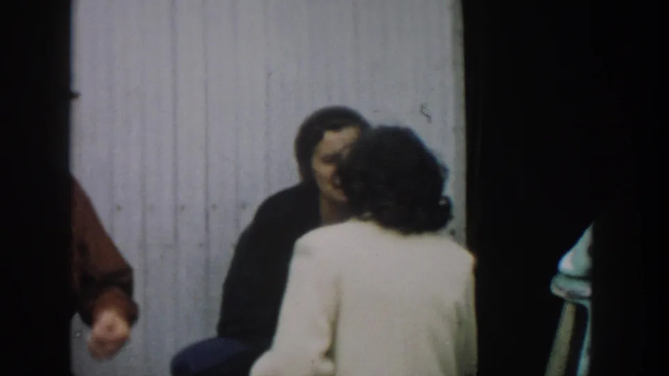 1957 Michigan home-movie frame with a woman in a white coat facing a smiling woman in a dark coat near an exterior doorway.