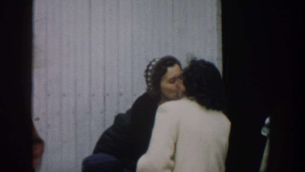 In 1957 Michigan home-movie footage, two women appear to embrace or speak closely beside a white corrugated wall.