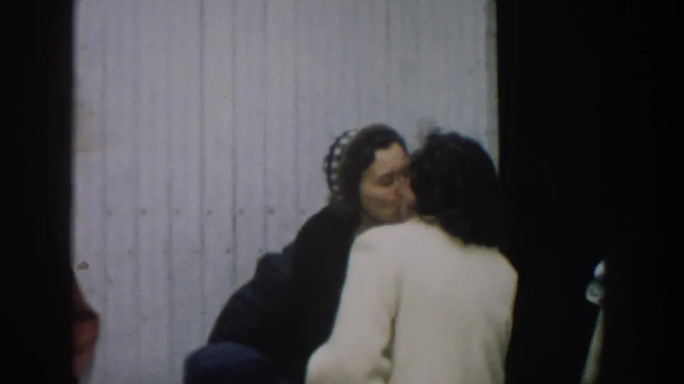 In 1957 Michigan home-movie footage, two women appear to embrace or speak closely beside a white corrugated wall.