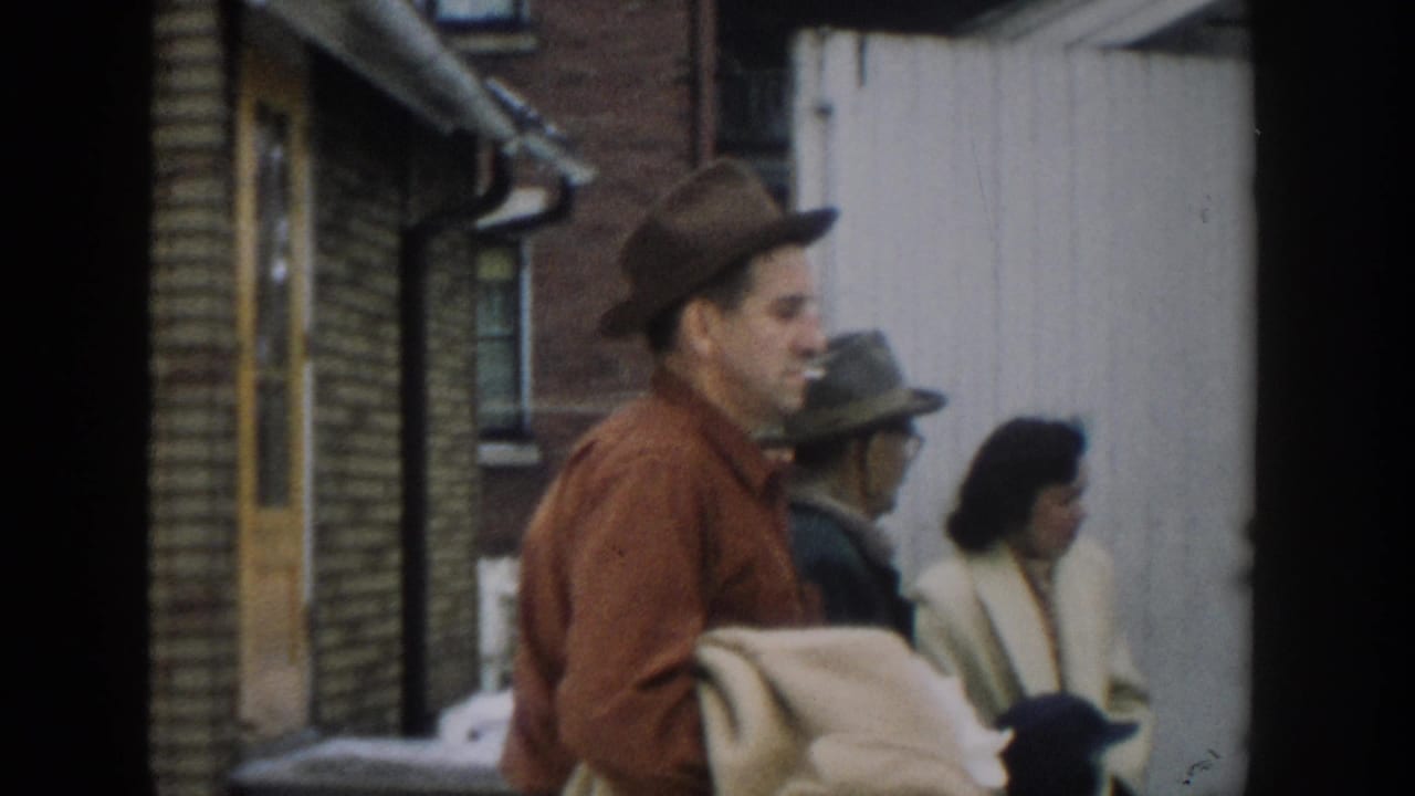 A man in a brown hat and red shirt moves through a Michigan street scene with other adults in period coats.