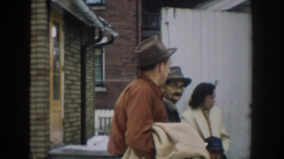 The home-movie frame shows adults in 1950s-style hats and coats gathered near brick and light exterior walls in Michigan.