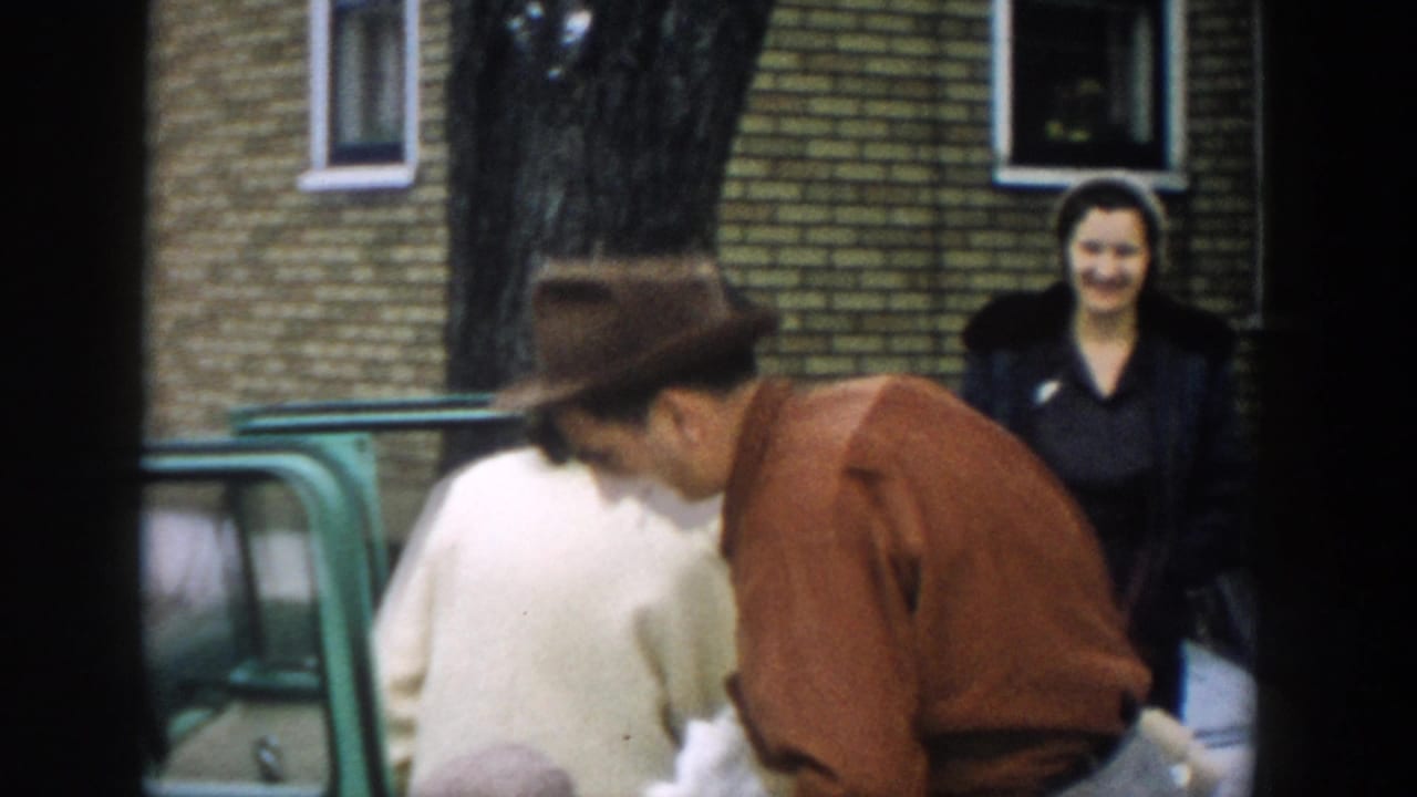 1957 Michigan home-movie frame showing a man in a brown fedora beside a green station wagon as women stand near a brick building.