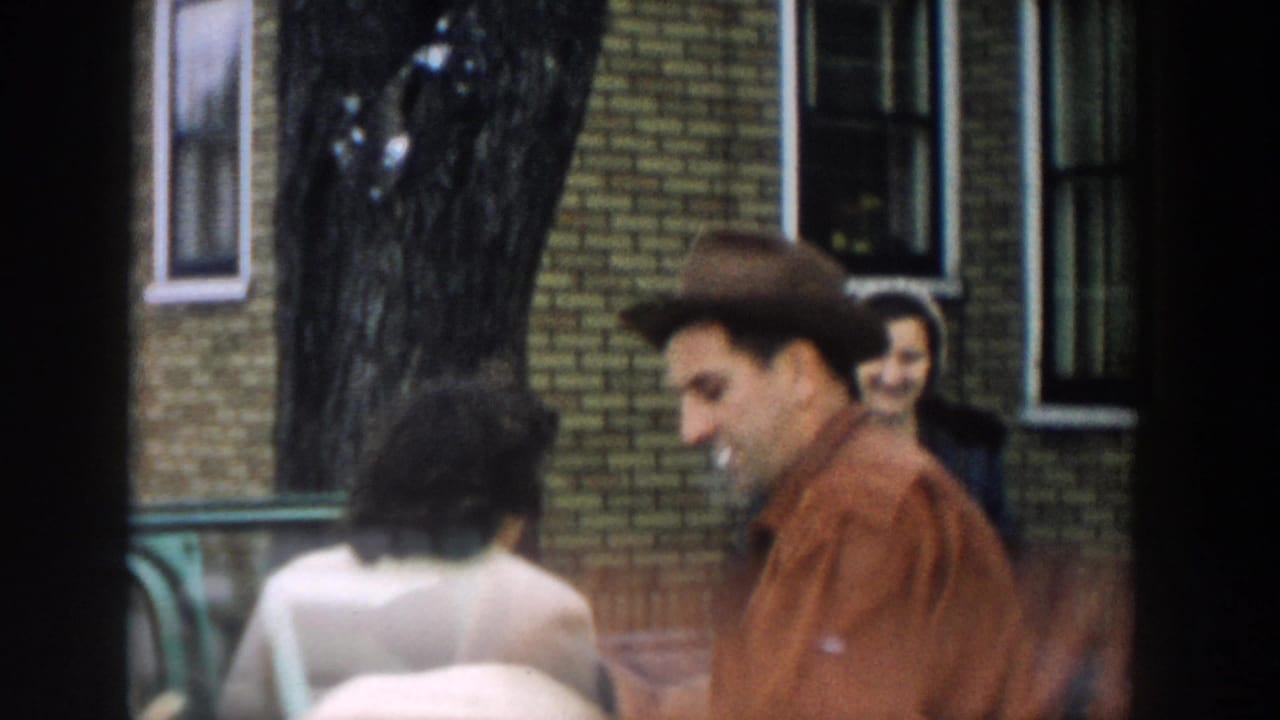 Man smoking near a green station wagon with women and a brick building behind him.