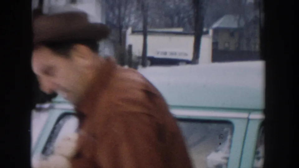 A blurred close pass of the man in brown beside the green station wagon, with snow and bare trees visible beyond the car.
