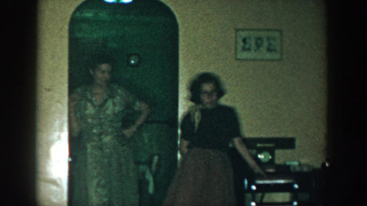 1957 Michigan home-movie frame showing two adult women in a dim private room near an arched doorway and small wall pictures.