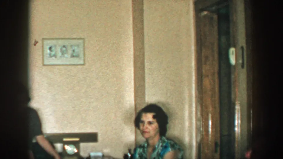 1957 Michigan interior frame with a seated woman near wood doors and a partly visible standing person in soft focus.