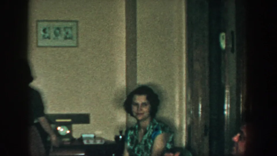 1957 Michigan home-movie frame showing a seated woman in a patterned top beside a wall fixture and wood door.
