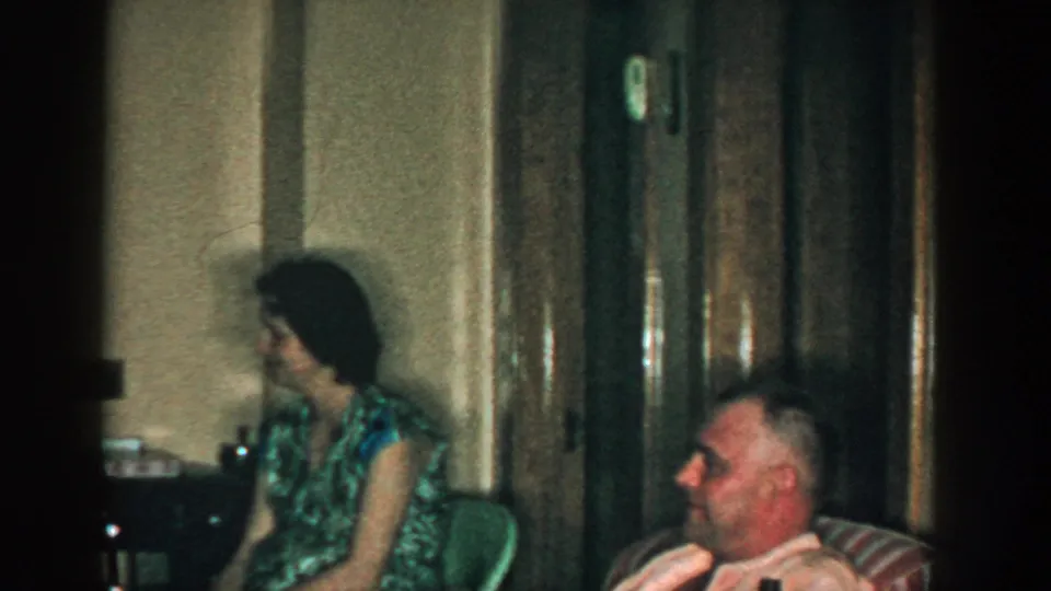 1957 Michigan frame showing a seated woman and seated man near wood doors during a quiet indoor gathering.