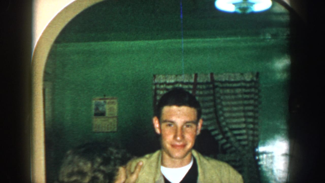 1957 Michigan home-movie frame showing a young man smiling in a dim green interior, with a curtain and arched opening behind him.