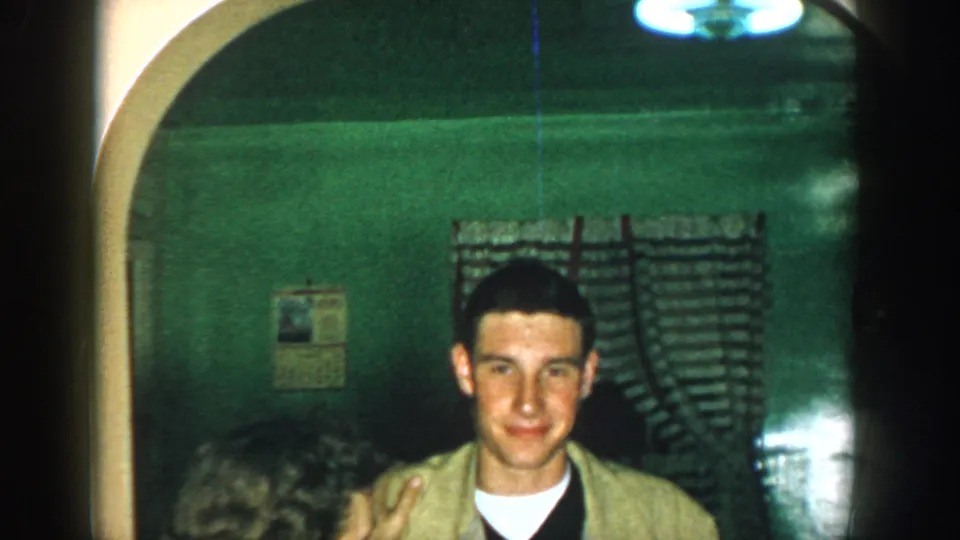 1957 Michigan home-movie frame showing a young man smiling in a dim green interior, with a curtain and arched opening behind him.