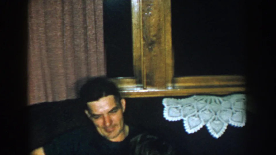 1957 Michigan home-movie frame showing the man seated on a couch by a curtained window and lace textile in a private room.