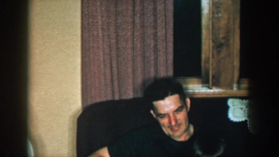 1957 Michigan home-movie frame with a seated man near a window and another person briefly entering the foreground.
