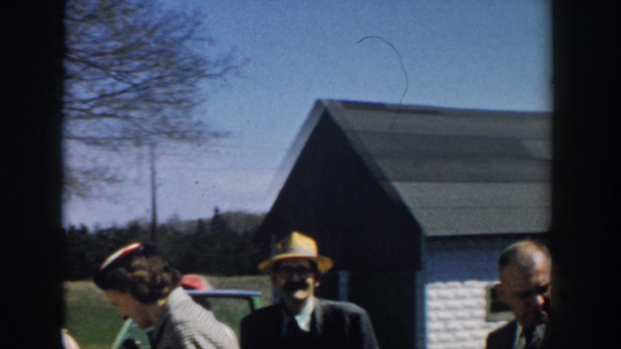 Adults in hats and suits stand near parked cars by a rural building and trees.