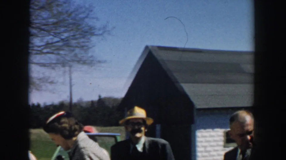 1957 Michigan 8mm frame showing adults gathered near mid-century cars, with a dark-roofed building and leafless trees behind them.