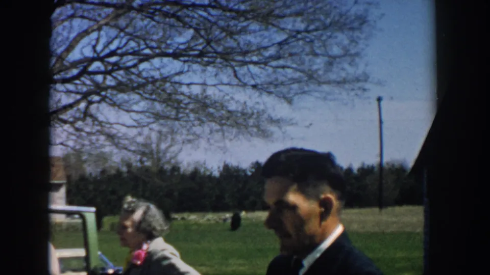 1957 Michigan home-movie frame showing a suited man in the foreground, an open car door, and adults in a rural field setting.