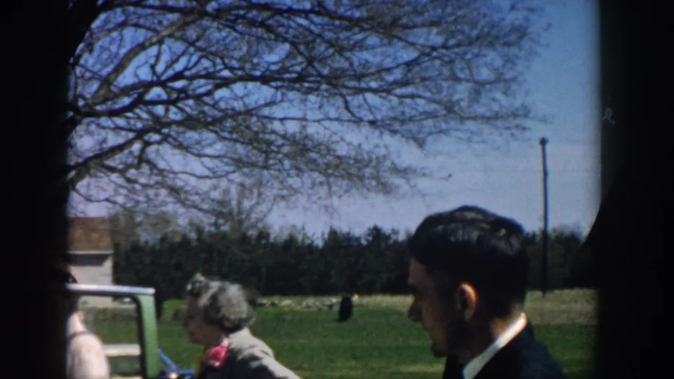 A 1957 Michigan 8mm frame with a suited man and gray-haired woman beside an open car door, framed by trees and open grass.