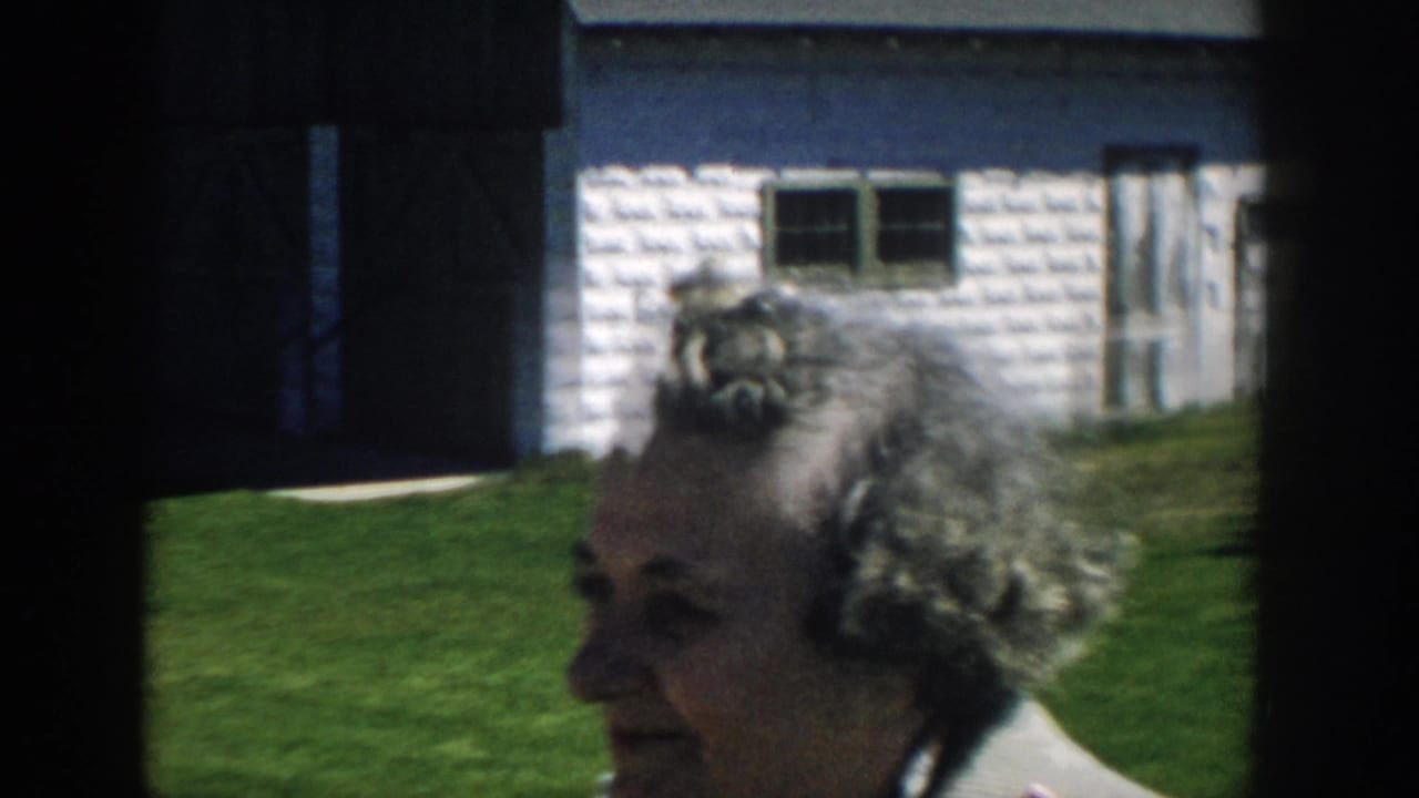 Older woman in profile outside a small white-sided house on a green lawn.