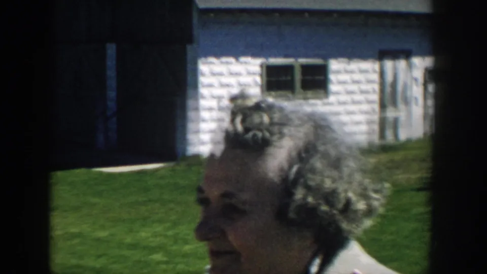 Older woman with gray styled hair seen in profile outside a small white-sided house in 1957 Michigan home-movie footage.