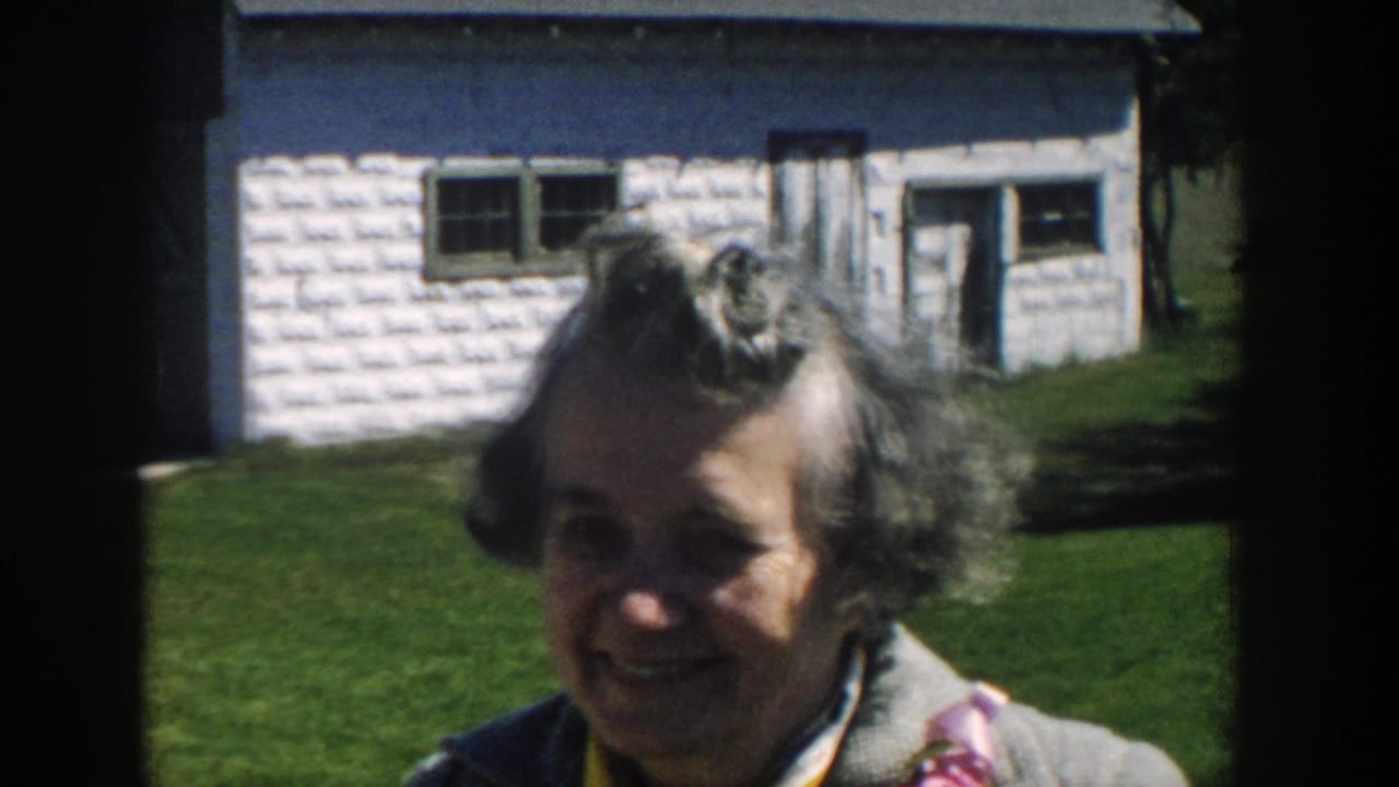 Older woman smiles toward the camera outside a small white-sided house in Michigan, supported by the 1957 clip metadata.