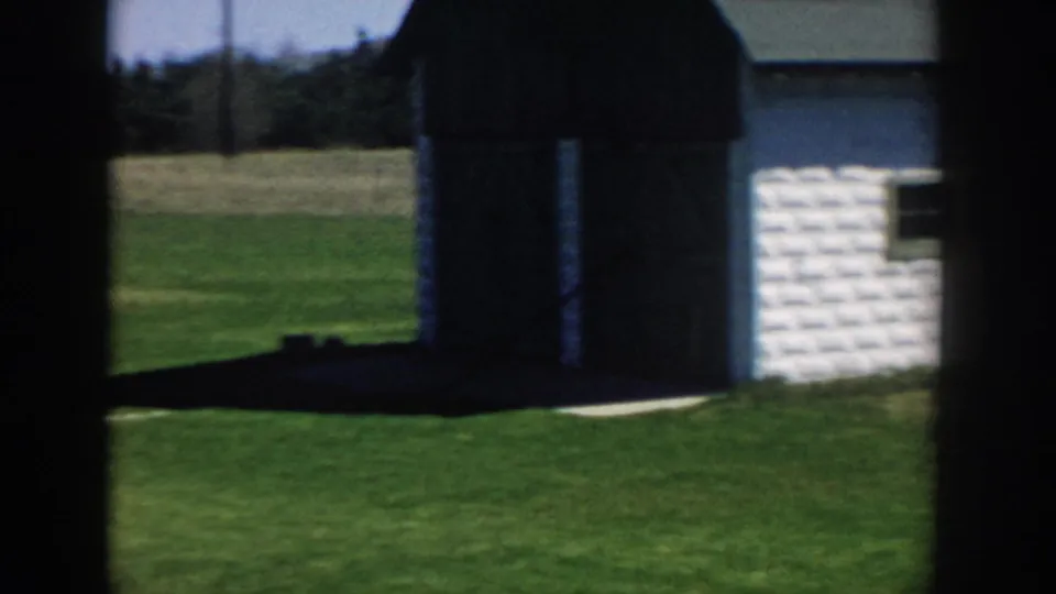 A Michigan rural yard appears with a white-sided building, green lawn, open field, and dark structure in 1957 home-movie footage.
