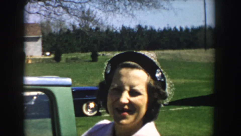 Woman in a dark hat stands near classic cars on a green lawn in the 1957 Michigan home-movie scene.
