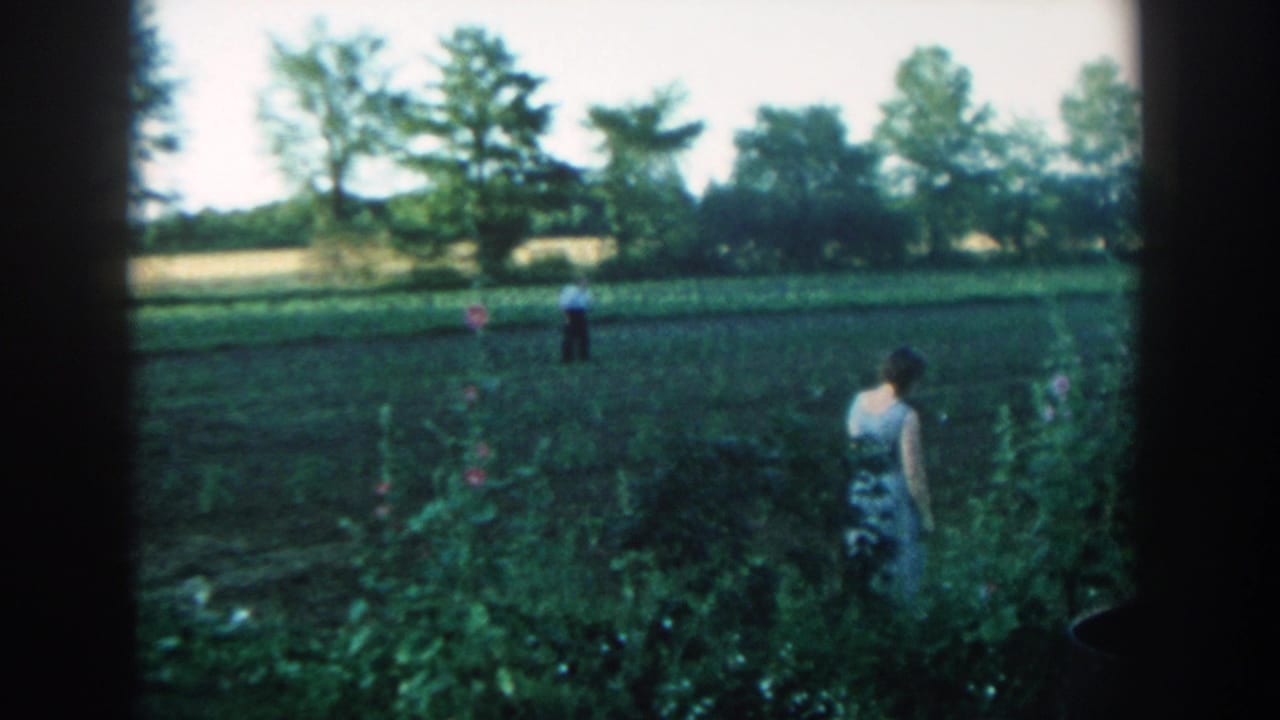 1957 Michigan 8mm frame with a woman near tall garden plants and a man standing at a distance across cultivated ground.