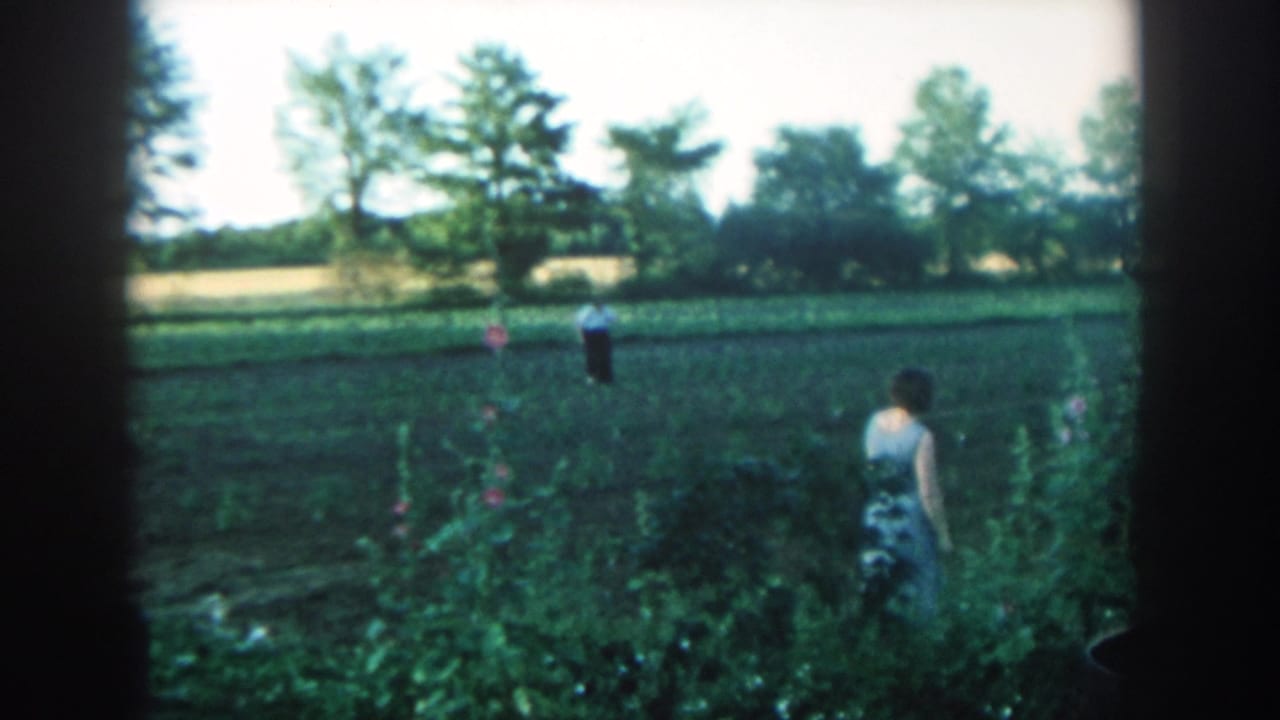 Woman faces away beside garden plants, with a man in a white shirt standing across the field.