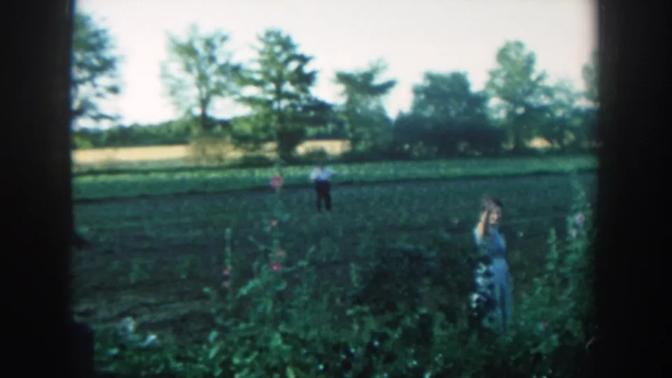 1957 Michigan 8mm still with the woman gesturing beside garden plants and the man standing farther back in the cultivated field.