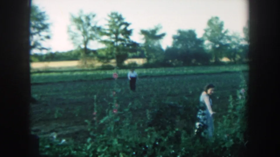 1957 Michigan home-movie frame with a woman turning beside dense garden plants and a man visible across the open field.