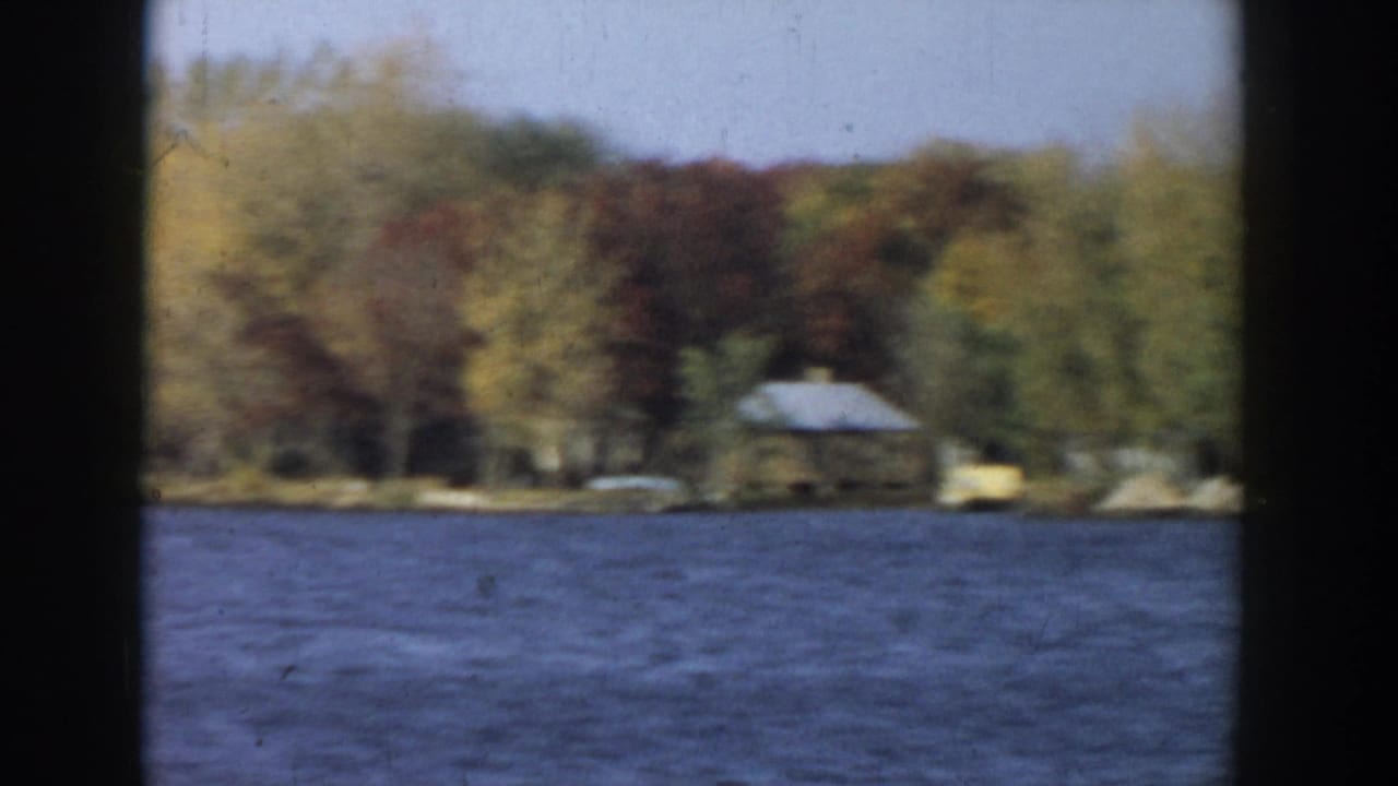 1957 Wisconsin lake view with rippled water, autumn trees, and a distant lakeside cabin, partly framed by dark window bars.