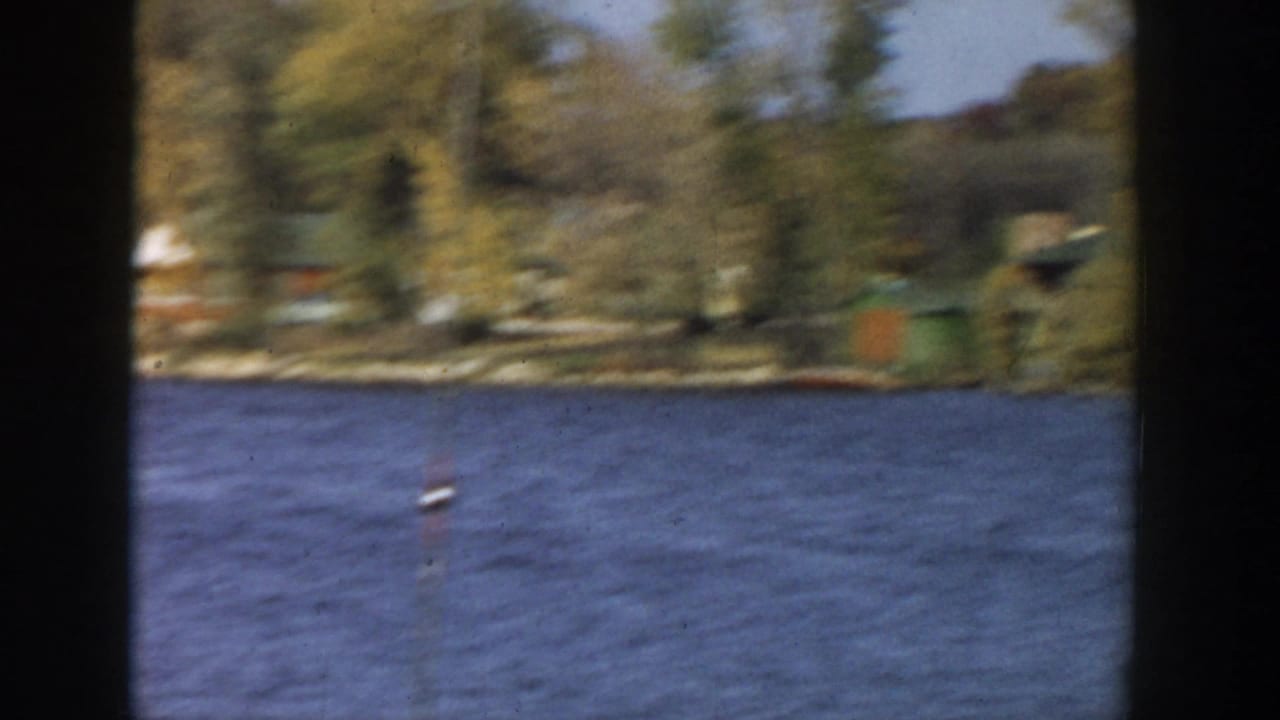 Soft 1957 Wisconsin 8mm frame of a lakeshore with cabins or small structures behind rippled water and autumn trees.