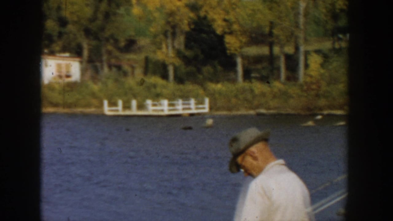 Man in a hat fishing near blue water with shoreline trees and white fencing in the background.