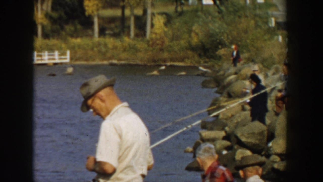 Several people fish from rocks along the water with a man in a hat in the foreground.