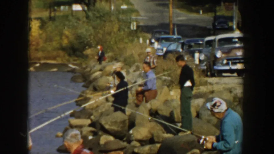 1957 Wisconsin home-movie frame showing anglers on rocks beside a lakeshore road with vintage cars behind them.