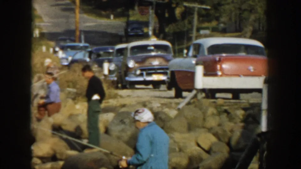 1957 Wisconsin lakeshore frame with people fishing from rocks and several late-1950s cars parked nearby.