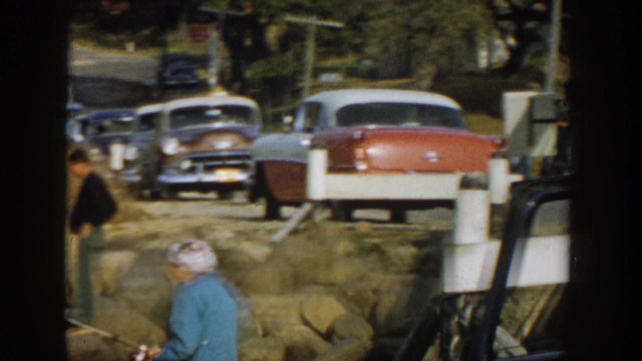 Roadside scene with parked vintage cars, two people near rocks, and white posts beside a road.
