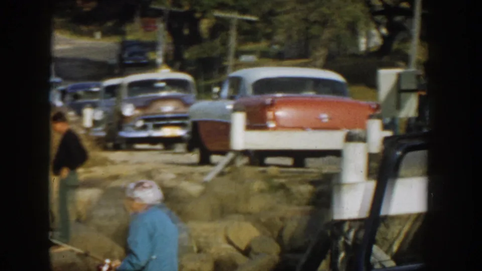 1957 Wisconsin home-movie frame showing parked mid-century cars and two people near a rocky roadside embankment.