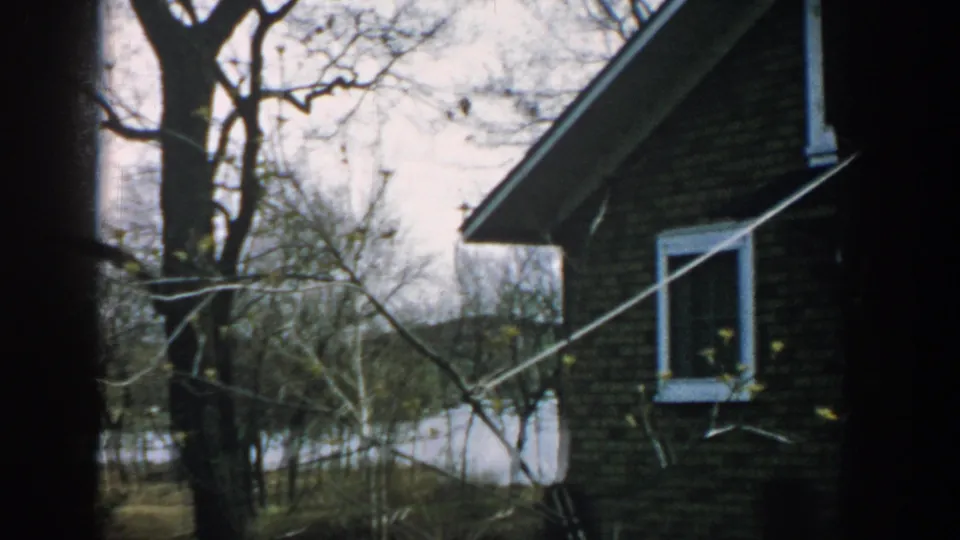 1957 Wisconsin 8mm frame of a brick house seen through bare trees, with white trim, yard, and water in the distance.