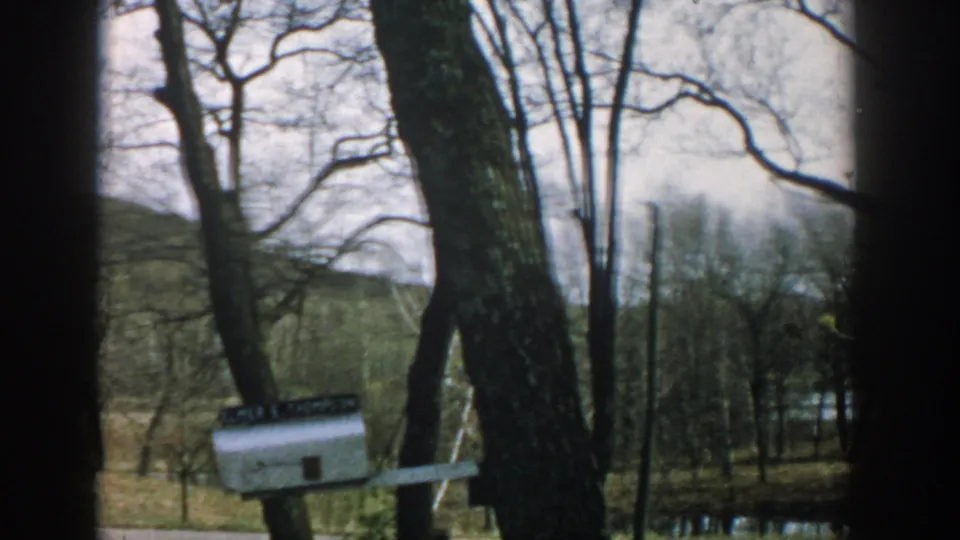 1957 Wisconsin home-movie frame showing a white mailbox among bare trees, with a house edge, yard, and water in the background.