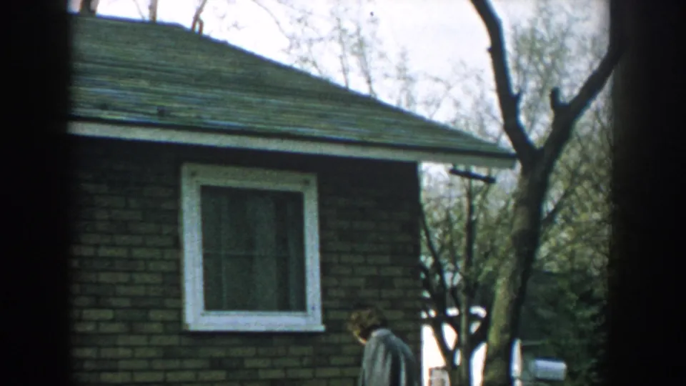 1957 Wisconsin 8mm frame with an adult man walking near a brick house, white-trimmed window, trees, water, and a mailbox.